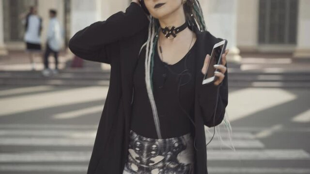 Unrecognizable millennial hippie dancing to music in headphones holding smartphone as friends chatting at background. Young confident Caucasian woman enjoying hobby in sunny summer city.