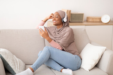 Joyful black Muslim woman in hijab and headphones listening to music on cellphone, closing her eyes in delight, at home