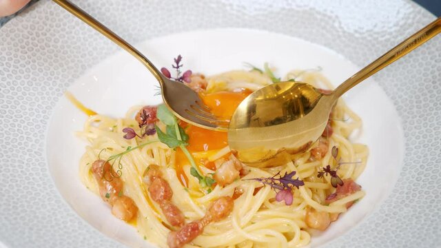 Close-up of delicious carbonara pasta with bacon and egg in a white plate. Hands taking spaghetti with cutlery.