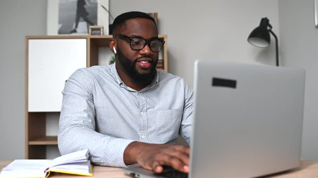 Cheerful young multiethnic guy in glasses using laptop sitting at the desk in home office. An African entrepreneur websurfing, answering email, chatting online with a smile