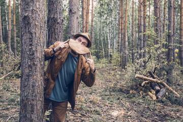 a grown man in a hat and jacket leans out from behind a tree and holds a segment of a log in front...