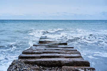 Fototapeta premium Waves break on rocks and fly apart into small drops and turn into foam. Long concrete breakwater stretching away into depths of stormy blue sea. Banner of sea landscape.