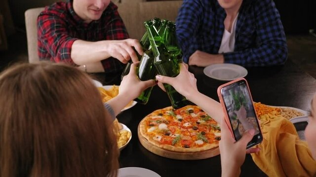 Friends clink bottles of beer and the girl takes pictures of it on the phone. A girl blogger takes pictures of pizza and food.
