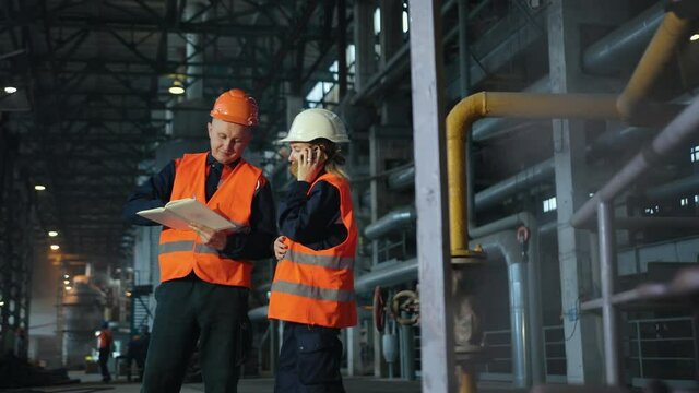 Two maintenance engineers man, woman inspect. Work heavy industry manufacturing factory. Mechanic, female engineer talking in plant. Young employees wearing helmets are working industrial factories