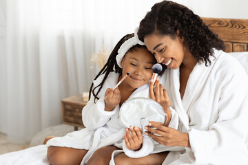 African mother and her little daughter in bathrobes making makeup on bed