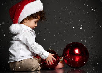 Naklejka premium Stylish handsome barefoot baby boy in Santa hat and trendy outfit playing xmas tree toy ball ornament under snowfall background. Wintertime, Christmas eve concept. Isolated portrait on gray copy space