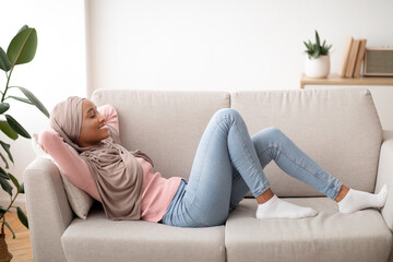 Beautiful black woman in hijab lying on sofa with closed eyes, taking break and relaxing at home, copy space