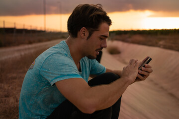 Young caucassian man with balbo beard wearing a blue t-shirt sitting outdoors at sunset and chatting with the smartphone  