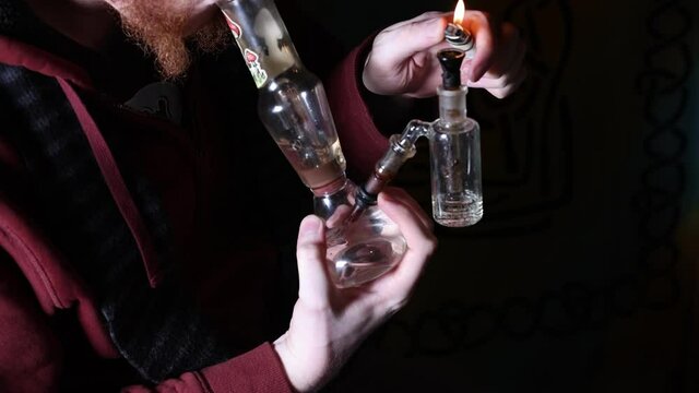 Bearded young male lights a bowl on a glass bong in slow motion.