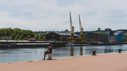 Un homme assit sur le quai de la seine à Rouen, en face de deux grues portuaires.