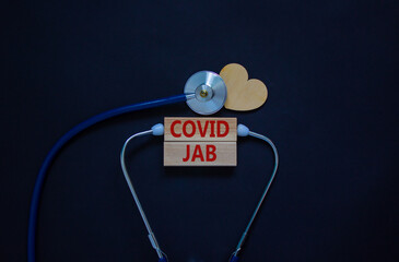 Covid jab symbol. Wooden blocks with words 'covid jab' and stethoscope on black background. Wooden...