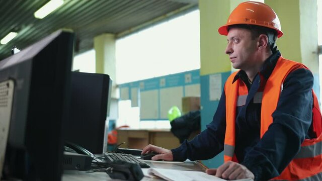 Inside heavy industry, factory male industrial engineer works on personal computer. Industrial engineer in hard hat wearing safety jacket uses computer. Works at heavy industry manufacturing factory