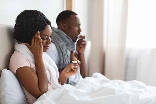 Sick black couple sitting in bed, measuring fever, sneezing noses
