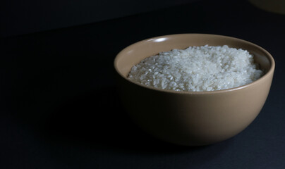 Fototapeta na wymiar White uncooked rice in a bowl with scattered grains nearby on a black background