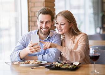 Cheerful millennial couple browsing internet on smartphone during romantic dinner at cozy cafe