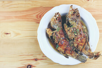 Thai Giant gourami fish and garlics deep fried and sweet spicy sauce in white dish on pine wooden table. Deep fried fish with Sweet chili. Thai food fried fish with spicy sauce. Thai style food.