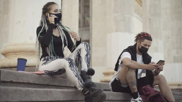 Young goth woman in Covid-19 face mask talking on the phone with boyfriend or friend surfing Internet on smartphone at background. Caucasian hippies on coronavirus pandemic outdoors.