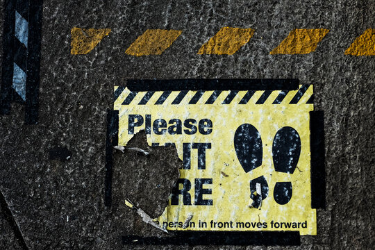 High angle close up of black and yellow 'Please Wait Here' sign on asphalt ground.