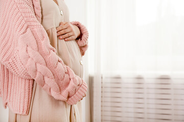 Cropped shot of young woman on second trimester of pregnancy. Close up of pregnant female in pink oversized knitted sweater with arms on her belly. Expecting a child concept. Background, copy space. - obrazy, fototapety, plakaty