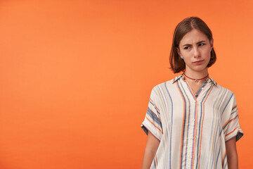 Young upset girl with necklace wearing striped shirt. Judgment thoughts about what happened. Watching to the left at copy space against orange background