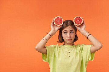 Young pretty woman with short brunette hair, looks sad, upset. Holding grapefruit over her head. Standing over orange background. Wearing green t-shirt, braces and bracelets