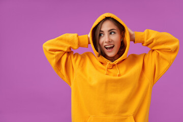 Teenage girl, cheerful and happy, with brunette short hair. Holding her head with hands and looking to the left at the copy space over purple background. Wearing orange hoodie, rings and teeth braces