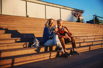 Cute happy senior couple in love sitting outdoors on the stairs and drinking coffee to go. Active seniors concept.