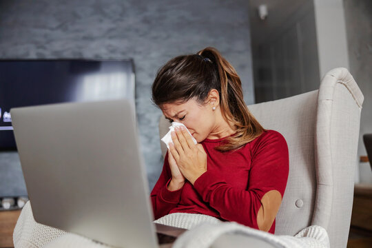 Ill young woman sitting at home, blowing her nose and holding laptop in lap. She is taking advices from doctors on iternet.