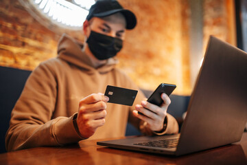 Shopping and online payment using cellphone, laptop and credit card. Man wearing black medical mask during Covid-19 sits in a cafe and buys goods via internet. New normal and technologies concept.