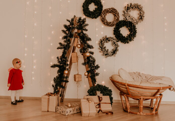 child stands at white wall with garlands Christmas tree with wrapped gifts