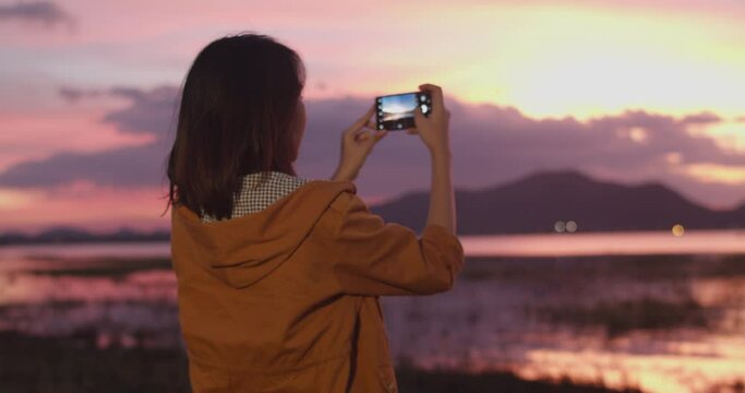 Cheerful young traveler Asian lady with backpack using phone for take a picture in camping on beach at night. Girl enjoy holidays adventure feeling happy freedom. Lifestyle travel and relax concept.