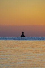 Naklejka na ściany i meble Vertical shot of a silhouette of a lighthouse in the middle of the sea at su