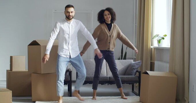 Young people two multinational people newlywed couple dancing actively moving to music make synchronized movements with hands and feet surrounded by boxes in new house, concept of buying real estate