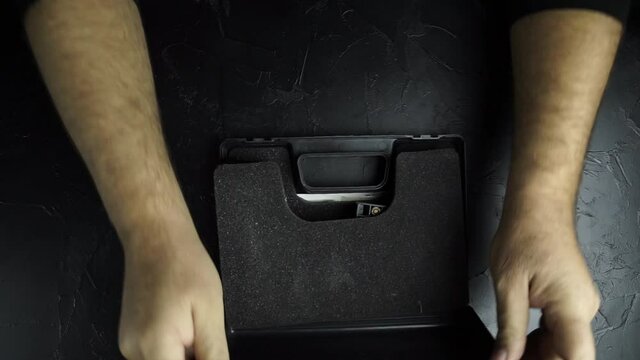 Top view of man's hands open black briefcase with firearms and ammunition, insert clip with bullets in pistol and distort bolt. Preparing 9-mm gun for shooting on texture table.