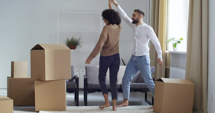 Multiethnic couple man and woman holding hands dancing whirling in living room next to cardboard boxes with things celebrating move to new home going to change apartment, happy family future concept