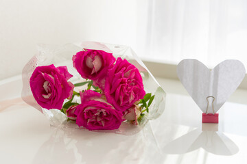 Roses on the table and a paper heart. Composition for Valentine's Day. A bouquet of delicate flowers in a package with a creative heart in bright sunlight. Layout of a gift card, paper valentine card