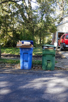 Overflowing green trash and blue recycling cans in a street by the curb on trash day near a driveway