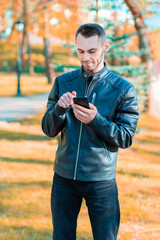 Youthful Guy Using Black Smartphone at the Beautiful Autumn Park. Handsome Young Man with Mobile Phone at Sunny Day - Medium Long Shot