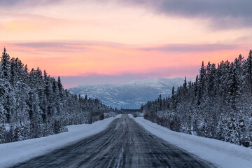 The Alaska Highway in Yukon Territory, northern Canada during December, winter time season with snow, snowy landscape on a cold, morning sunrise with pink, purple, orange colors. 