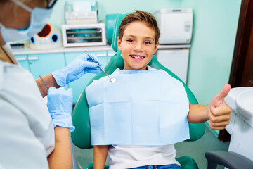 Small, young patient boy is happy with a visit to the dentist. Concept of painless dental treatment. Beautiful, wide smile of a child sitting in a dental chair in a doctor's office. Thumbs up