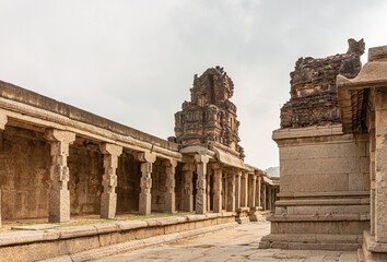 Obraz premium Hampi, Karnataka, India - November 5, 2013: Sri Krishna temple in ruins. Brown stone demolished back of side entrance Gopuram under silver sky and standing on beige stone pillared hall along outside w