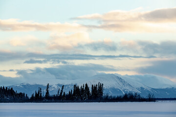Snow capped mountains in Yukon Territory, northern Canada with woods, forest and frozen lake in foreground during winter time season in December. 