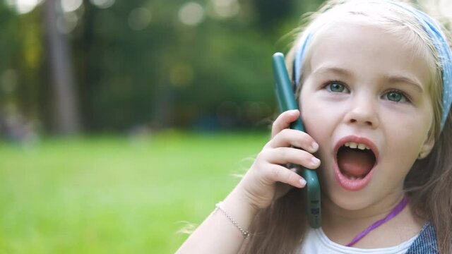 Happy little girl having conversation talking on mobile phone in summer park.