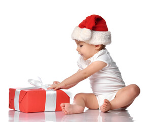 Cute infant baby girl wearing overalls, a red Santa Claus hat, sitting on a white floor studio, looking at a festive gift box wrapped with a bow. Isolated portrait. Christmas, New Year concept