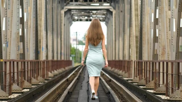 Back view of a slim woman with long hair walking along train tracks.