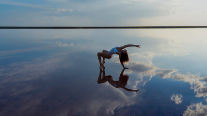 flexible slender girl gymnast performs dance posing in the reflection of the sky. salt lake, dancer against a dramatic sky with clouds