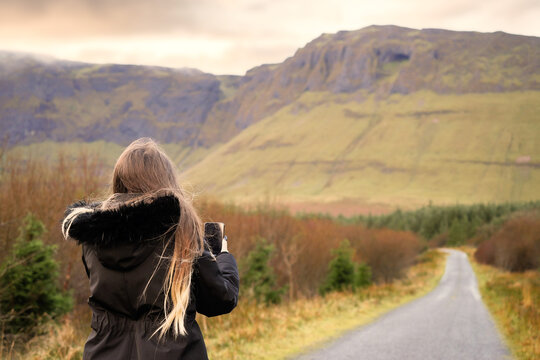 Teenager girl in focus taking picture on her smart phone of beautiful landscape. Gleniff horseshoe loop drive, county Sligo, Ireland.