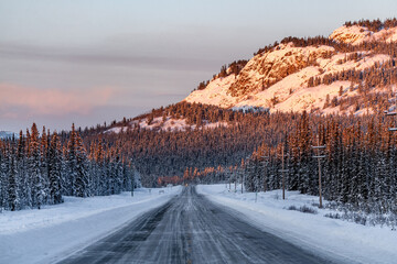 Obraz premium The snowy Alaska Highway in northern Canada, during winter time season in Yukon Territory. Sun shining on side of mountain with blue, clouds sky. 