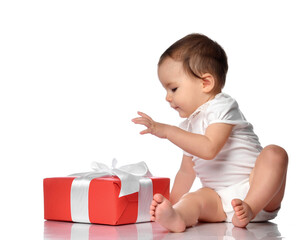 Baby and wrapped festive gift box with bow. Cute interested curious infant girl child in jumpsuit sitting on white floor studio background looking at present. Birthday or Christmas celebration concept
