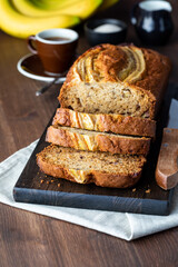 A close up of a sliced banana bread loaf on a wooden board with coffee and bananas in behind.